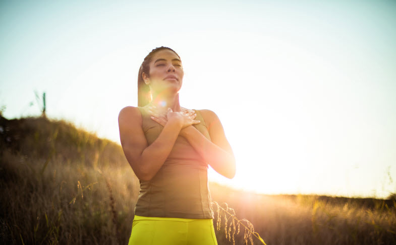 A woman standing outdoors in a field at sunset with her hands placed over her heart, eyes closed, and a serene expression on her face, embracing a moment of mindfulness and gratitude.