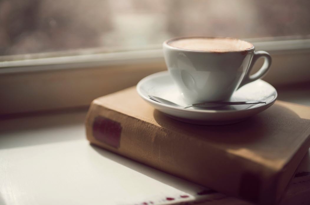 A cozy scene with a cup of coffee on a saucer, resting on top of a closed book near a window, suggesting a calm morning ritual.