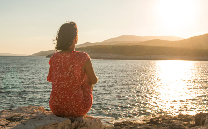 A woman sitting on a rocky shore, looking out over the water as the sun sets, conveying a peaceful moment of self-reflection.