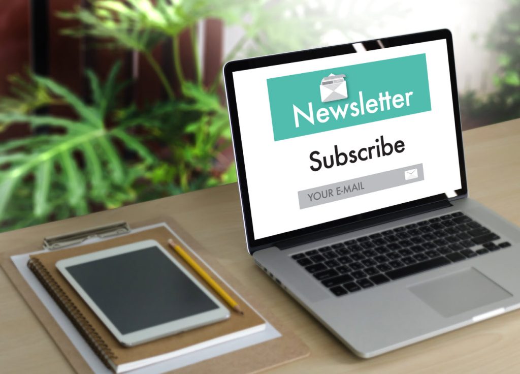 A laptop on a desk displaying a subscription page for a newsletter. Nearby are a notepad, tablet, and pencil, suggesting productivity and connection through digital communication.