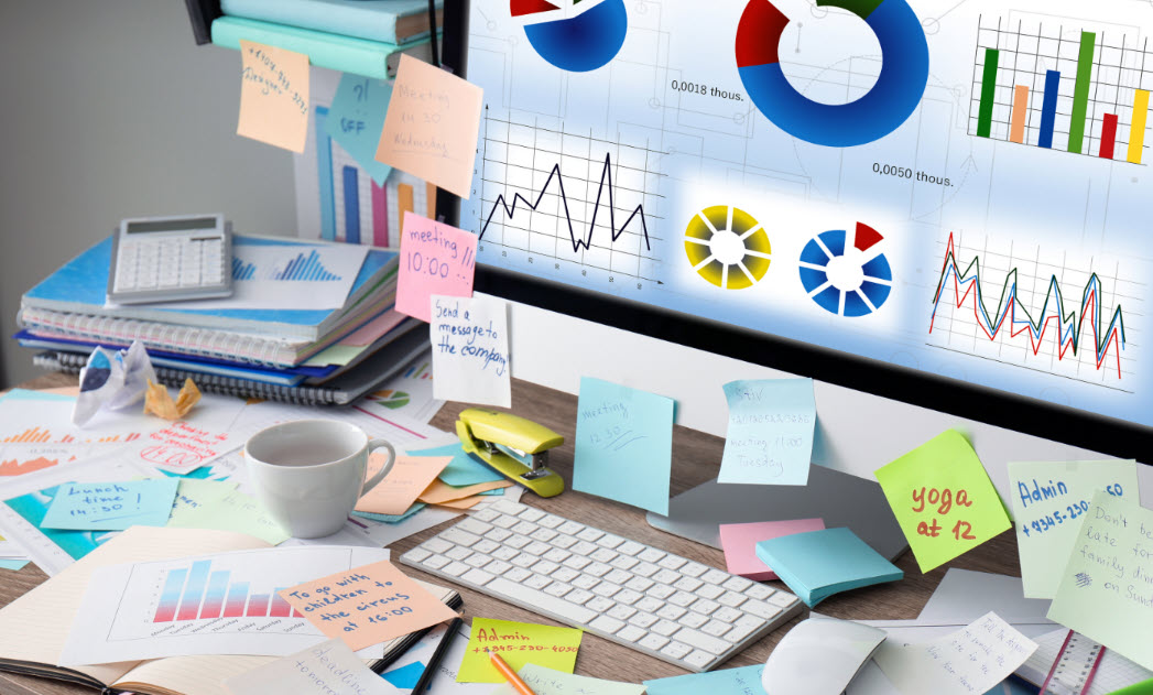 A desktop scene of post em' notes, spiral binders, coffee cup, monitor all indicating productivity and overwhelm in the normal course of a work day.