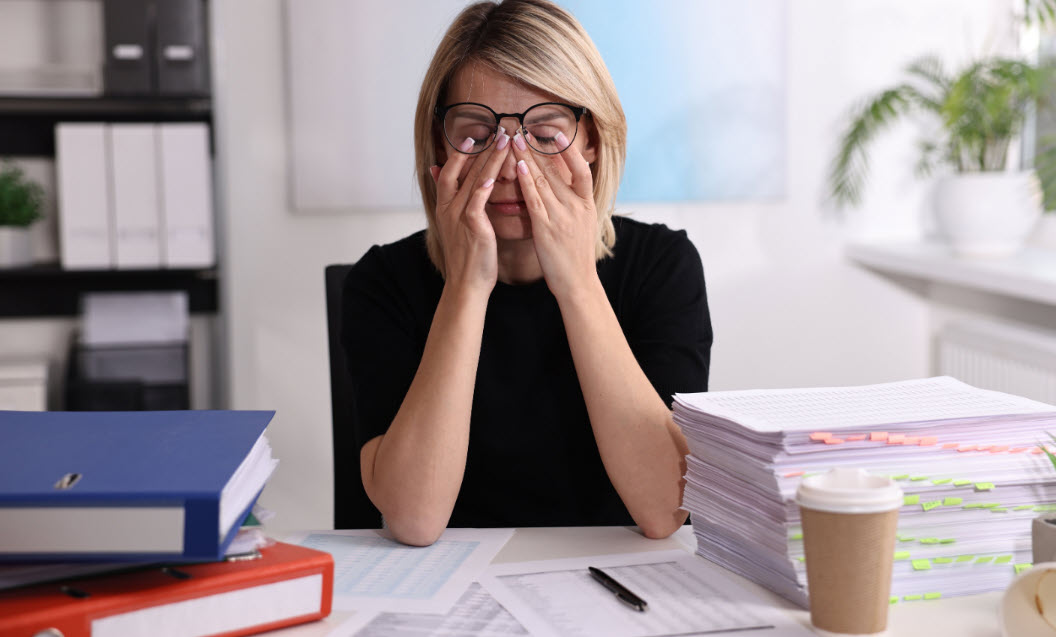 A woman sitting at her desk piled with a three reams of tagged sheets of paper to one side and folders on the other. The woman looks overwhelmed.