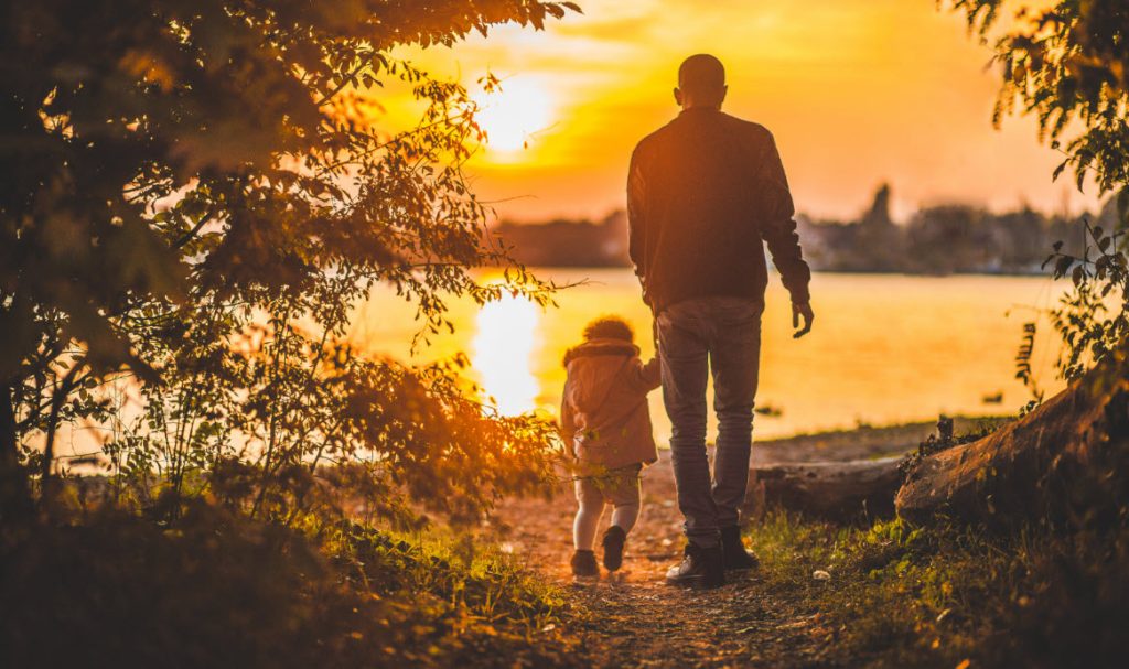 A scene where a father is bonding as he walks with his child towards the lake with the sun setting casting a golden hue. It's about teaching children early about nature and life.