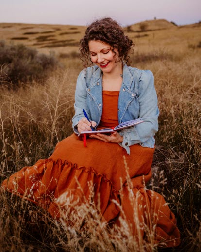 A woman with a burnt-orange dress and jean jacket is smiling as she sits in a hillside field. She appears to be free of any burdens as she's doing what fills her soul.