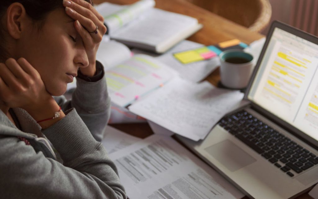 A young woman cradles her head with her hands. Before her are papers, charts and a laptop awaits while she takes a few minutes to pause and breathe from overwhelm.