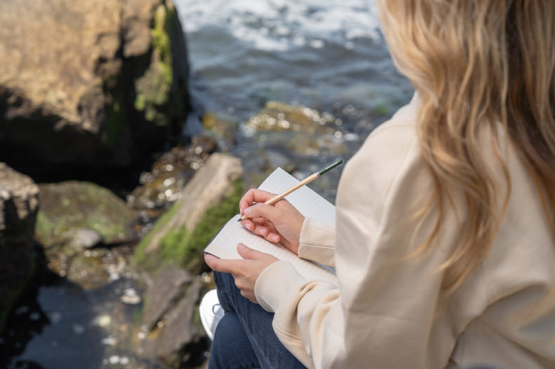 A woman sits atop a boulder looking down at boulders below her as the ocean laps gently against the wall of boulders and rocks. Create space for yourself, and listen to your inner wisdom for purpose.