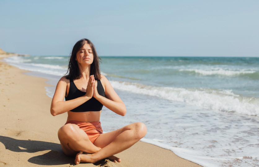A woman sits meditating next to shore where small waves lap near her, embodying focus and tranquility.