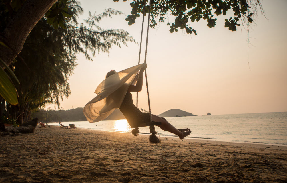 A woman swings on a beach side tree-hung swing. Her scarf softly trailing behind her. The sun is setting behind the distant hill leaving a soft shades of orange hue in the sky. Truly a micro-moment of presence.