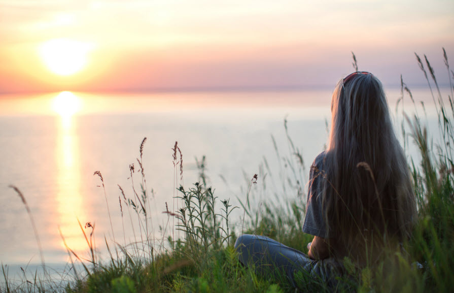 A woman sitting on a hill overlooking a lake with the sun setting on the horizon. What if purpose could simply mean living in alignment with your soul—whether or not it aligns with the blueprint the world has written for you?