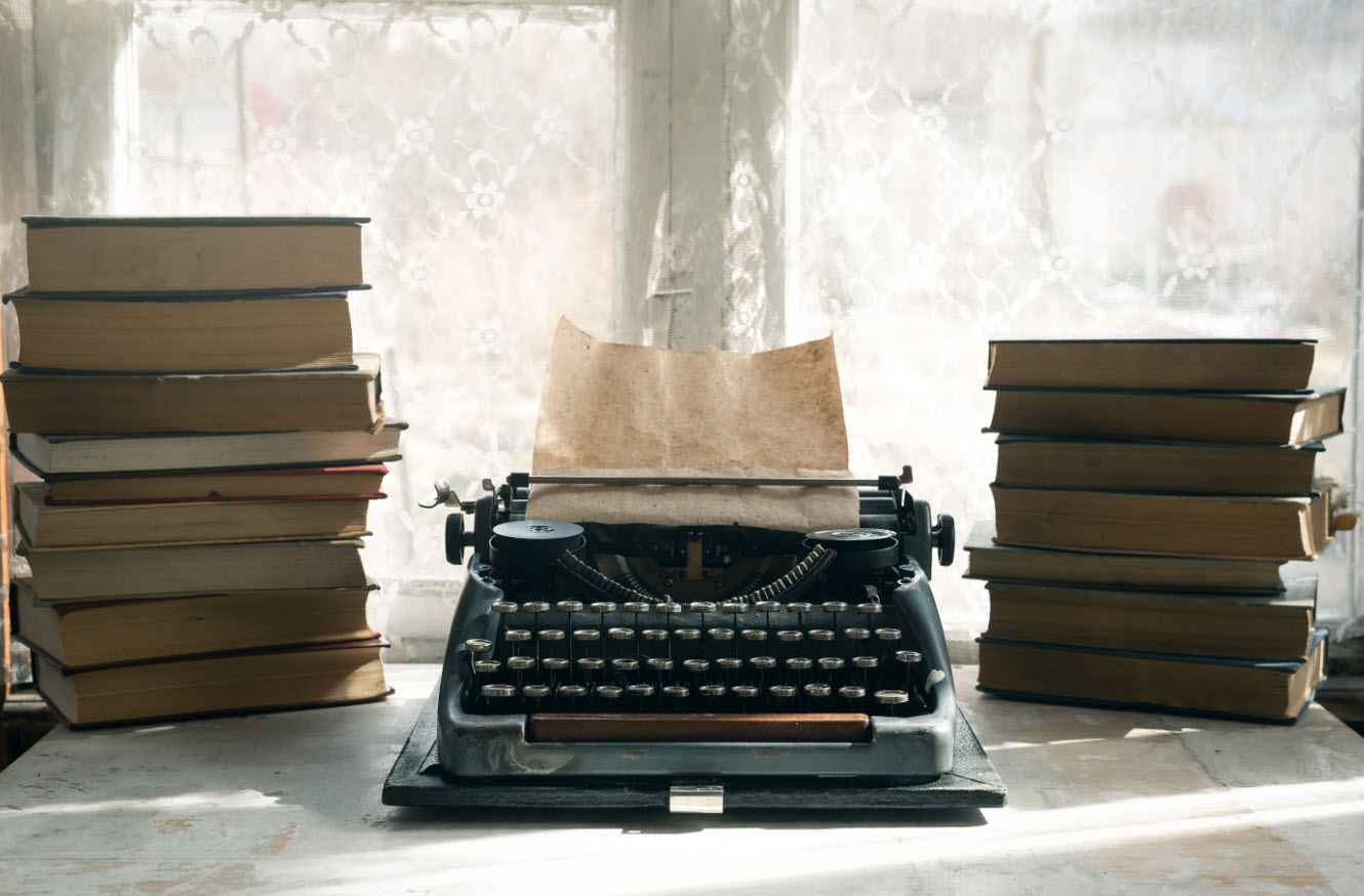 An antique typewriter with brown paper in its carriage sits centered on a table top. Along each side stands a stack of old hardcover books. Is purpose really tied to productivity or the approval of others? Re-write your purpose.